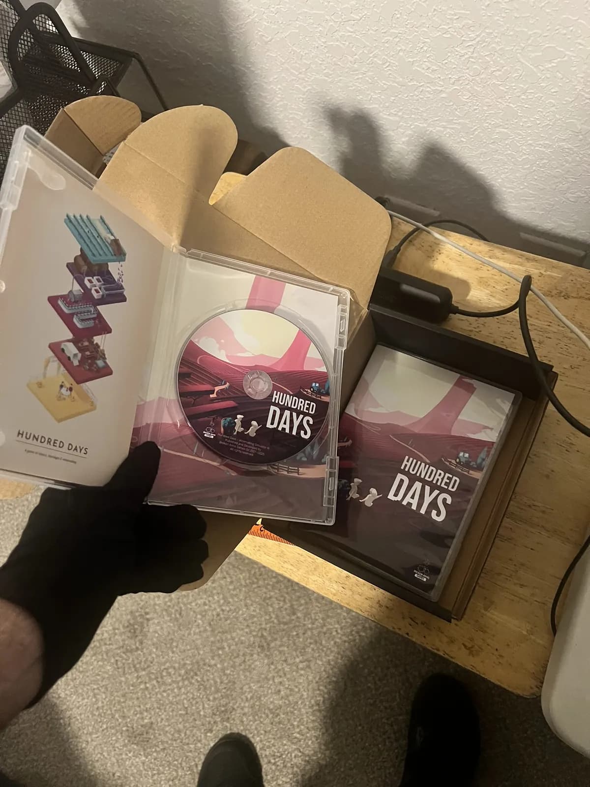 An open Hundred Days case showing the printed disc and interior art, held above a shipping box of finished copies.