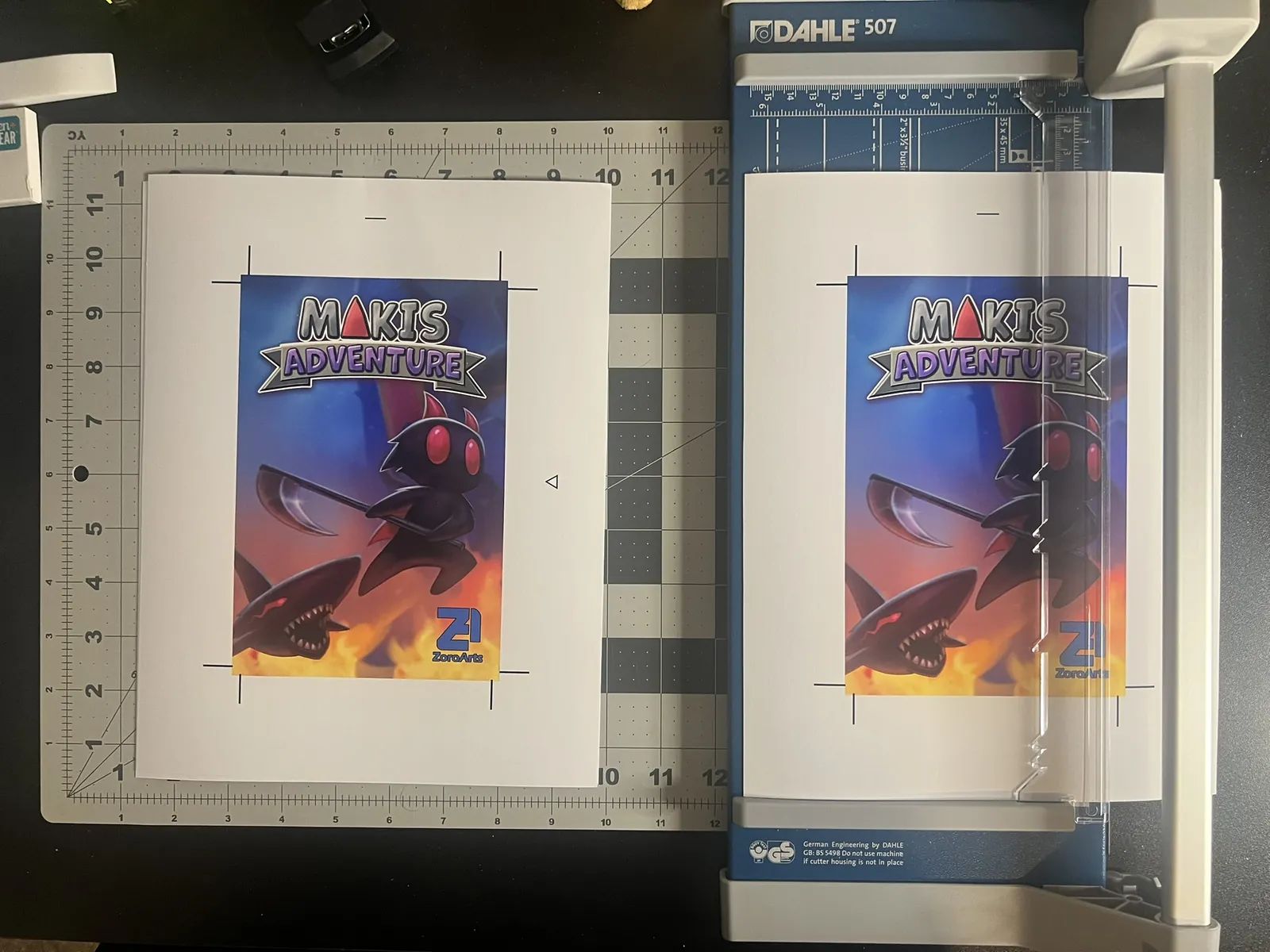 Two printed Maki's Adventure covers on the workbench: one already trimmed on a cutting mat, one still on the trimmer.