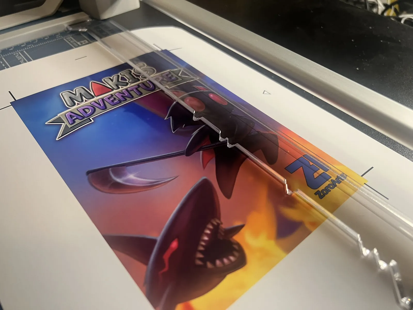A printed Maki's Adventure cover held flat under a rotary trimmer's guard rail, crop marks visible at the corners.